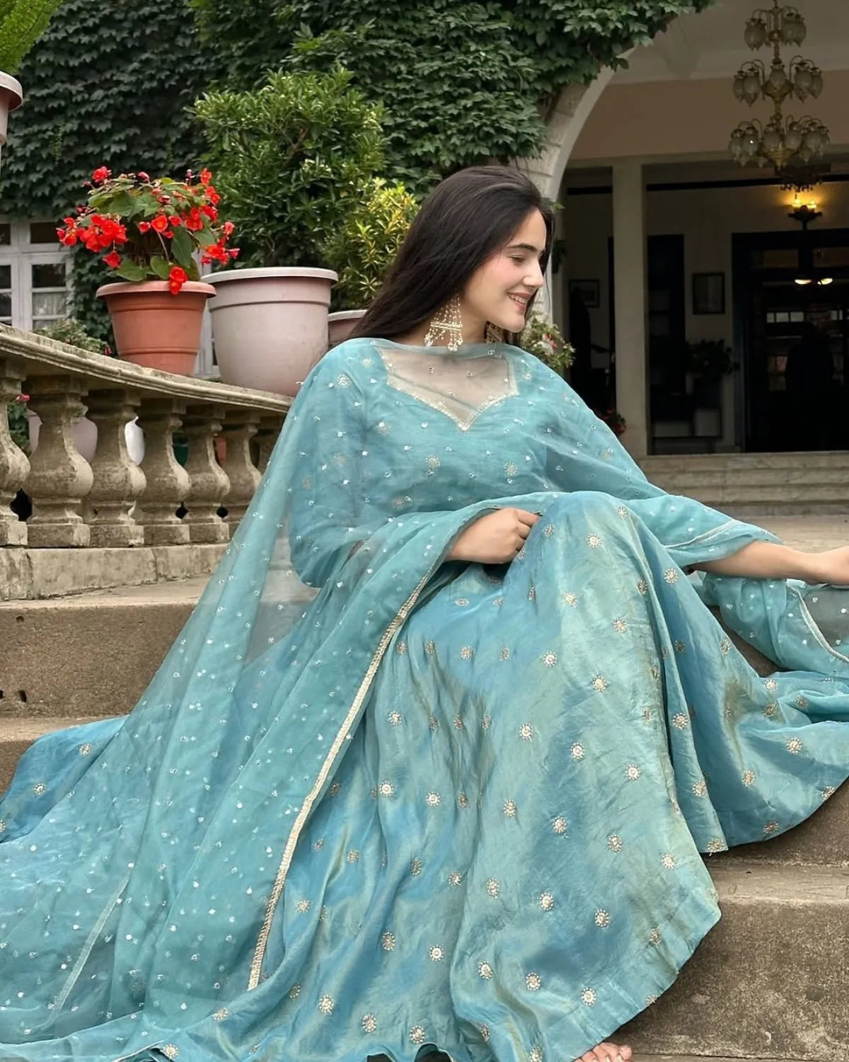Woman in a light blue saree sitting outdoors with plants and a building in the background
