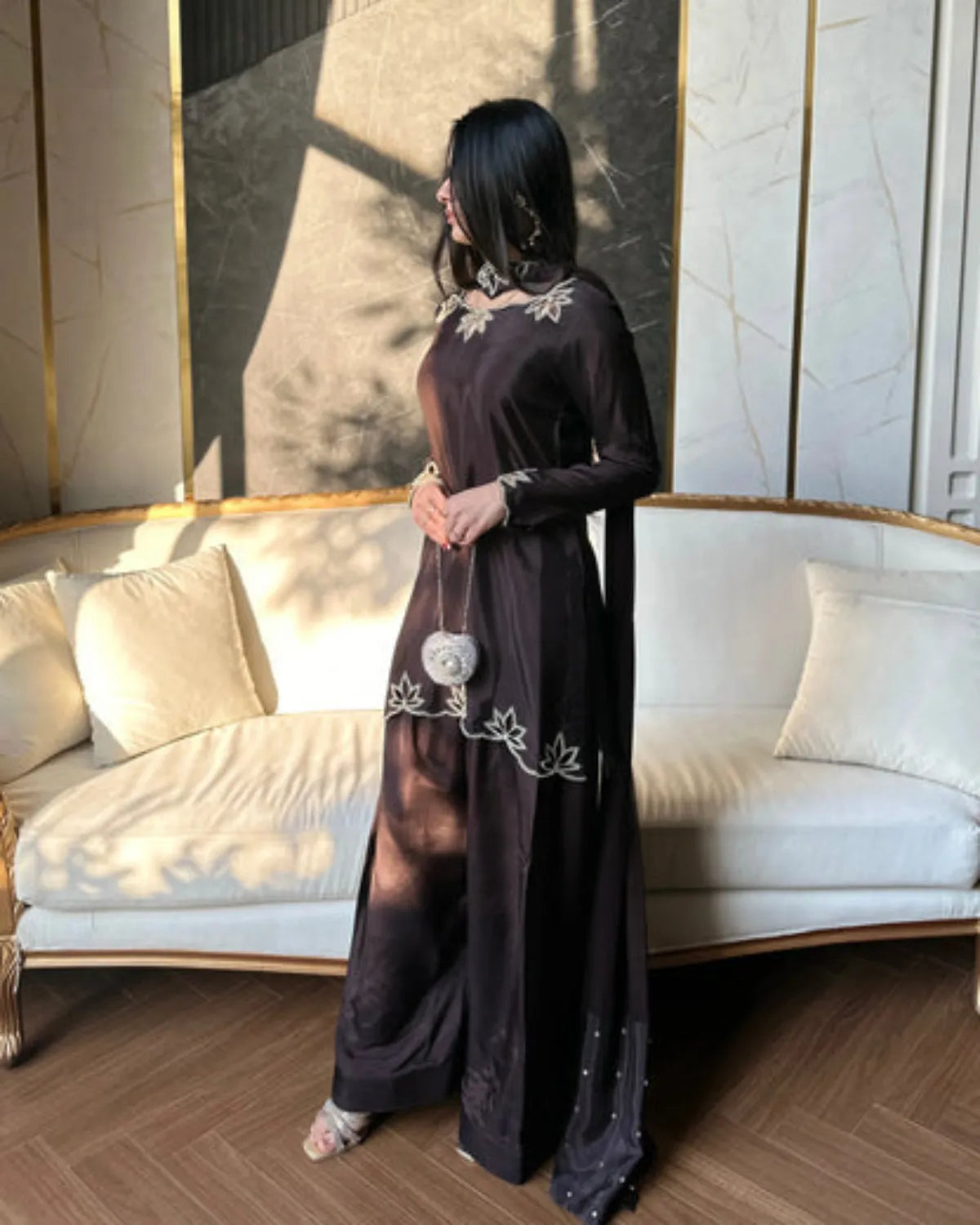 Woman in a long black dress with white embroidery standing in a stylish room.