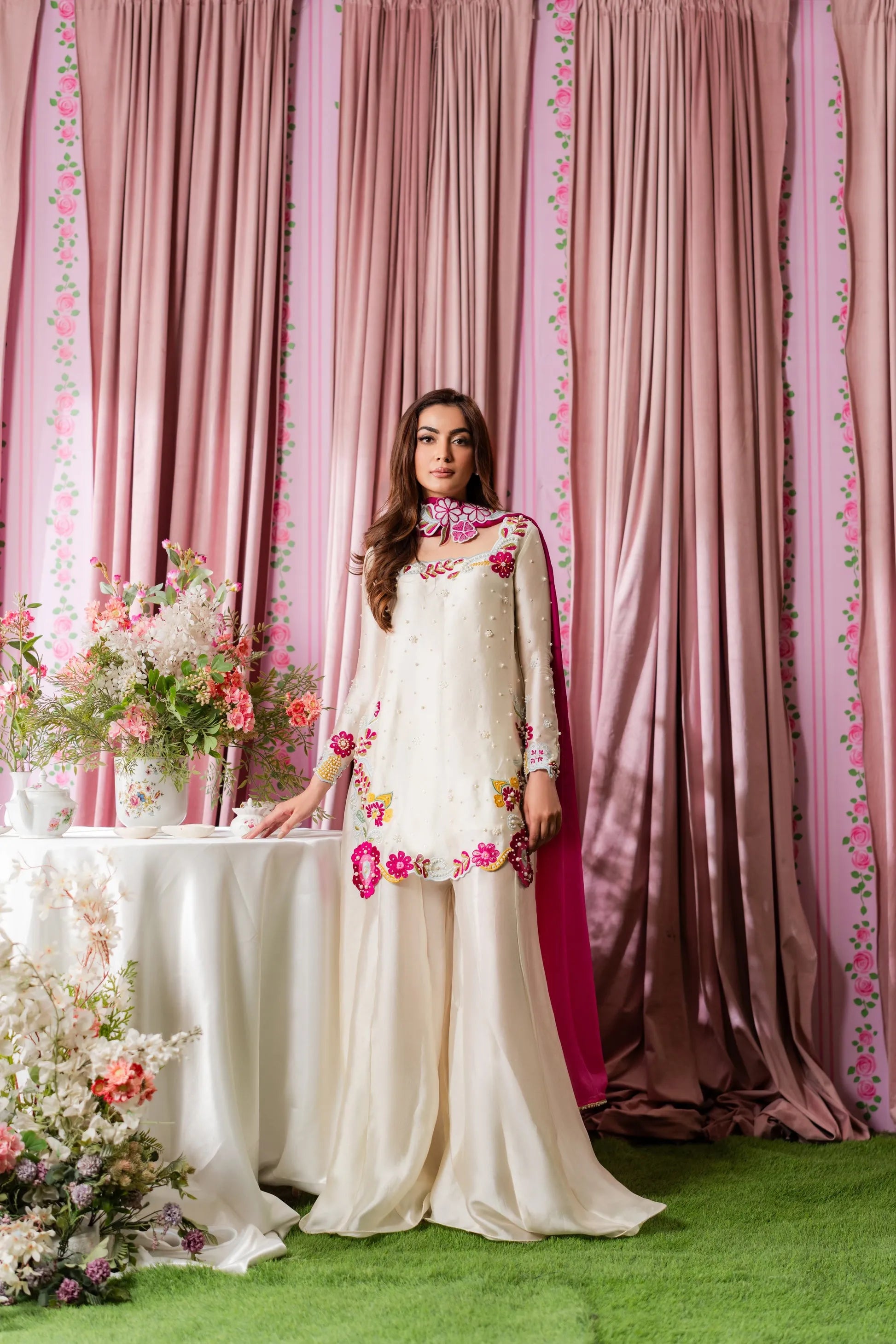 Woman in a white dress with floral patterns standing in front of pink curtains.