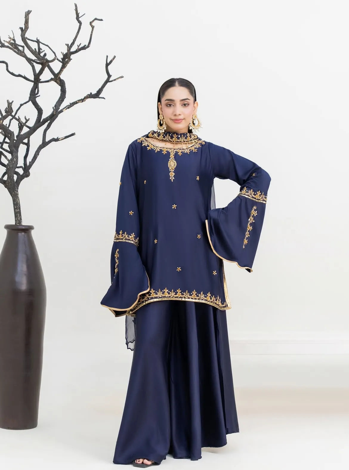 Woman wearing a navy blue traditional outfit with gold embroidery, standing next to a vase on a white background.
