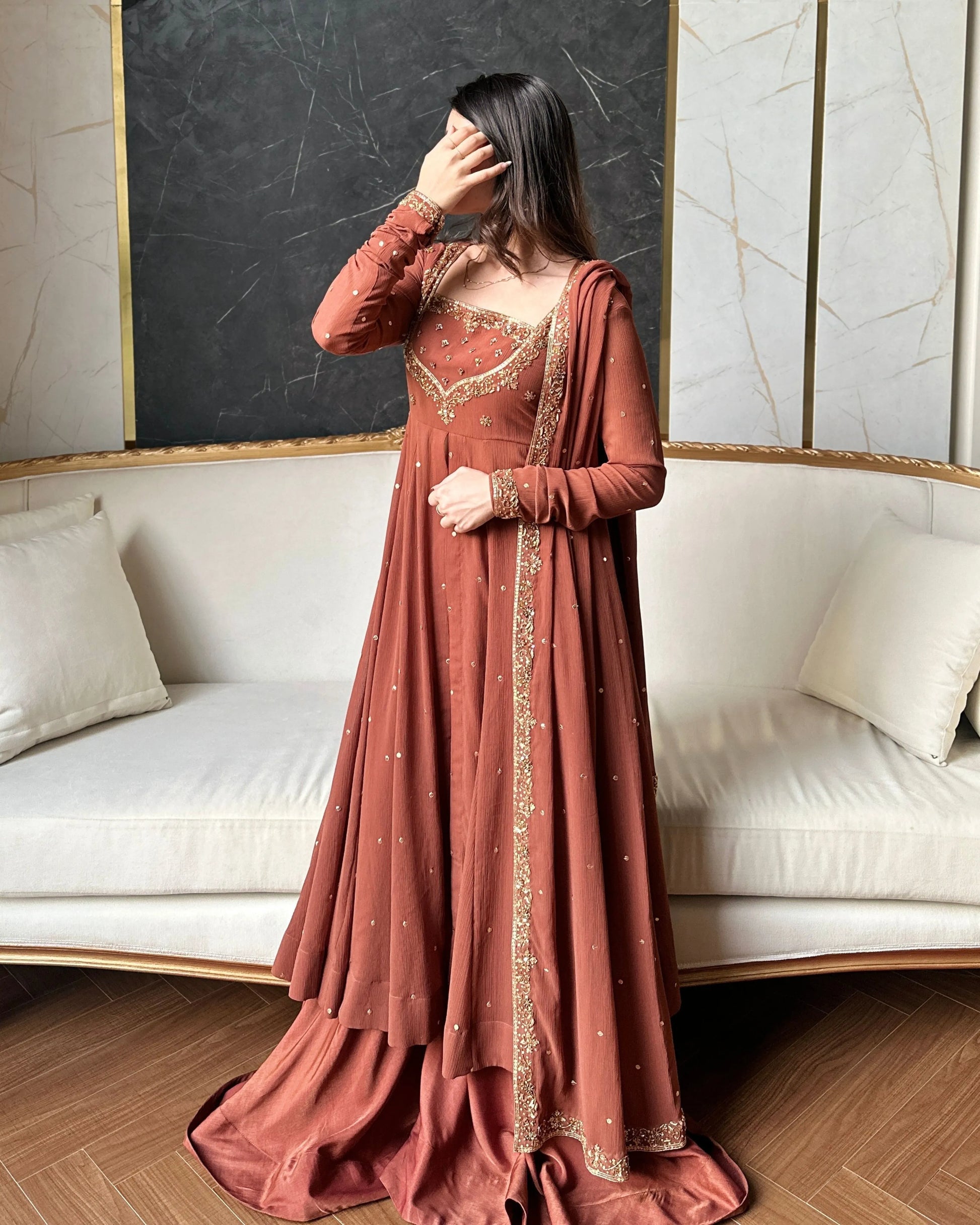 Woman in a rust-colored traditional outfit with gold embroidery standing in a room with a white couch and blackboard wall.