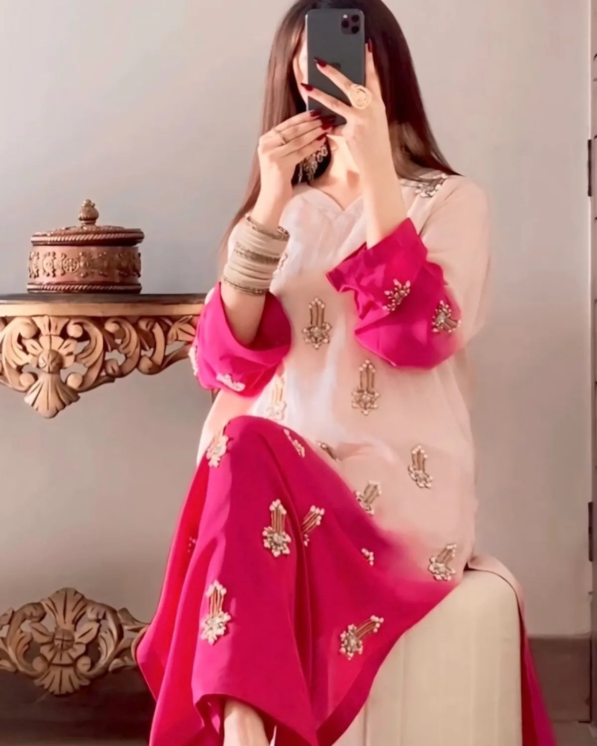 Person wearing a pink and white traditional outfit with gold embroidery, sitting on a couch.