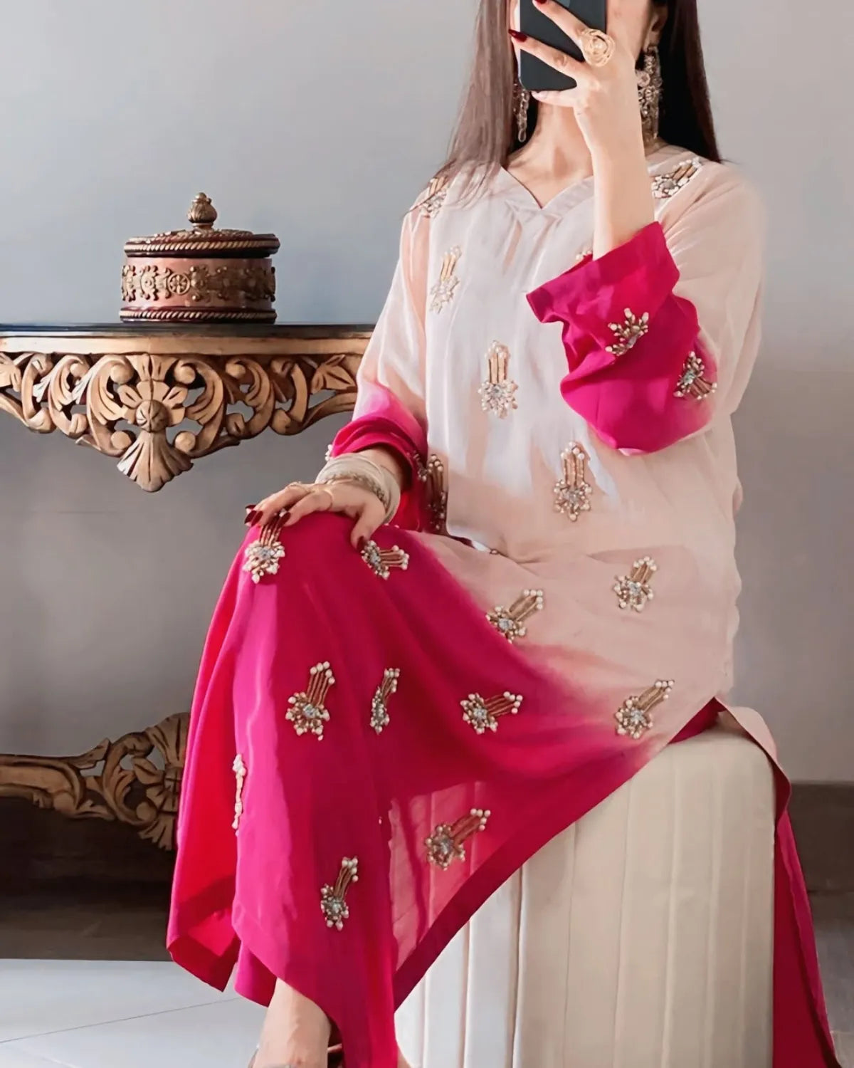 Person wearing a pink and beige saree with gold embroidery, sitting on a chair.