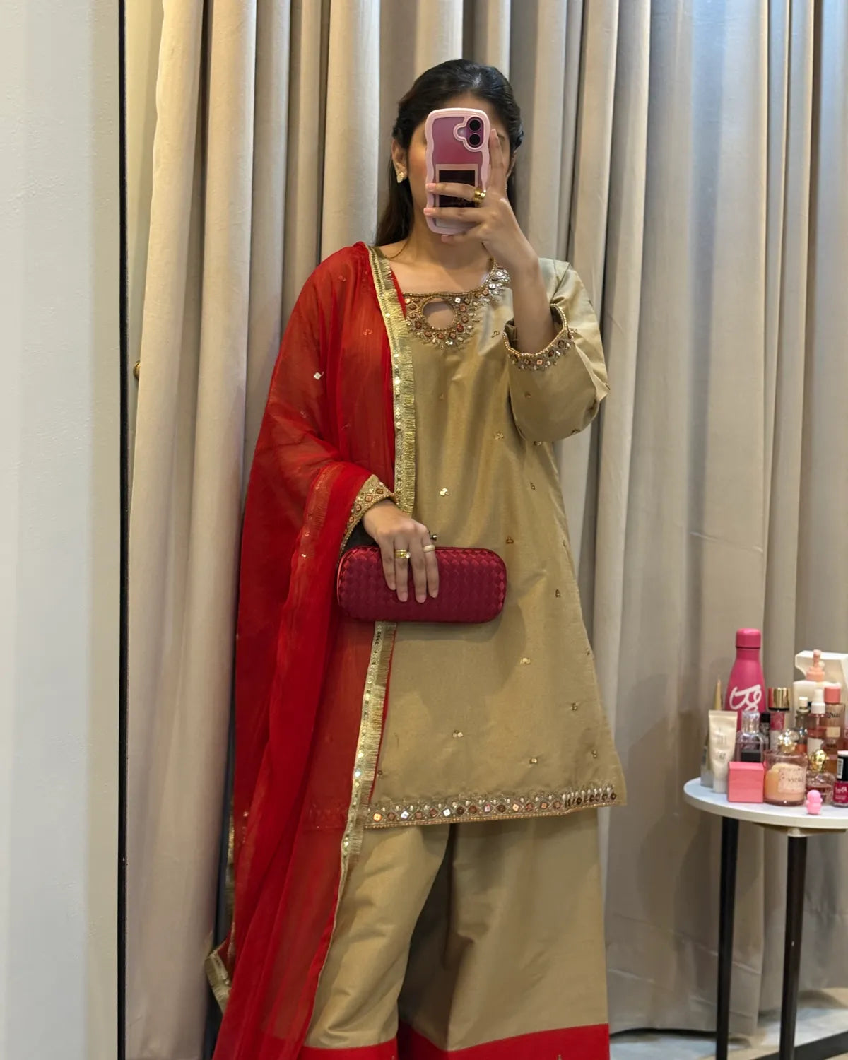 A woman takes a mirror selfie wearing a beige and red traditional Indian outfit with gold embroidery, holding a red clutch bag. The background shows beige curtains and a small table with cosmetics.