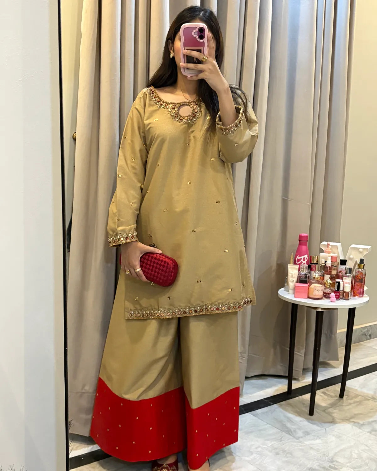 A woman takes a mirror selfie wearing a beige and red Indian ethnic outfit, consisting of a long-sleeve tunic with gold embroidery and matching wide-leg trousers with a red bottom hem. She is standing in front of beige curtains with a small table of cosmetics on the side.