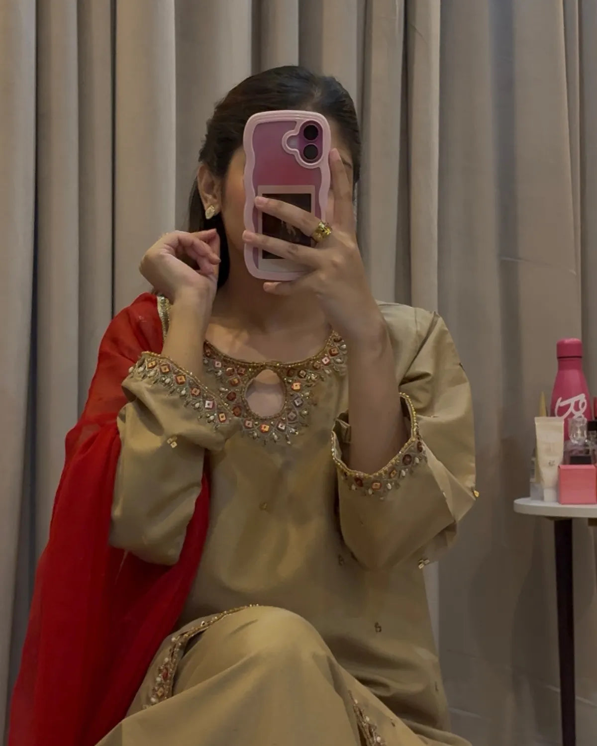A woman takes a mirror selfie wearing a beige embroidered traditional outfit with a bright red dupatta draped over her shoulder. The outfit features intricate gold and silver embellishments around the neckline and sleeve cuffs.
