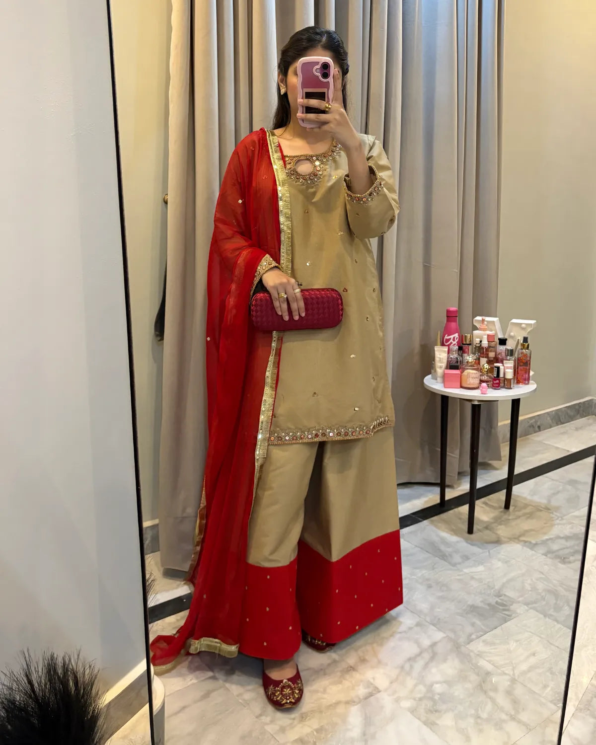 A woman takes a mirror selfie wearing a beige and red traditional South Asian outfit with a red dupatta, holding a pink phone. The setting is a fitting room with gray curtains and a small side table with cosmetics.