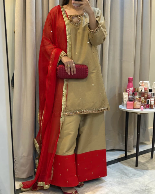 A woman wearing a beige and red traditional South Asian outfit with gold embroidery, holding a red clutch bag, standing in front of a mirror in a fitting room.