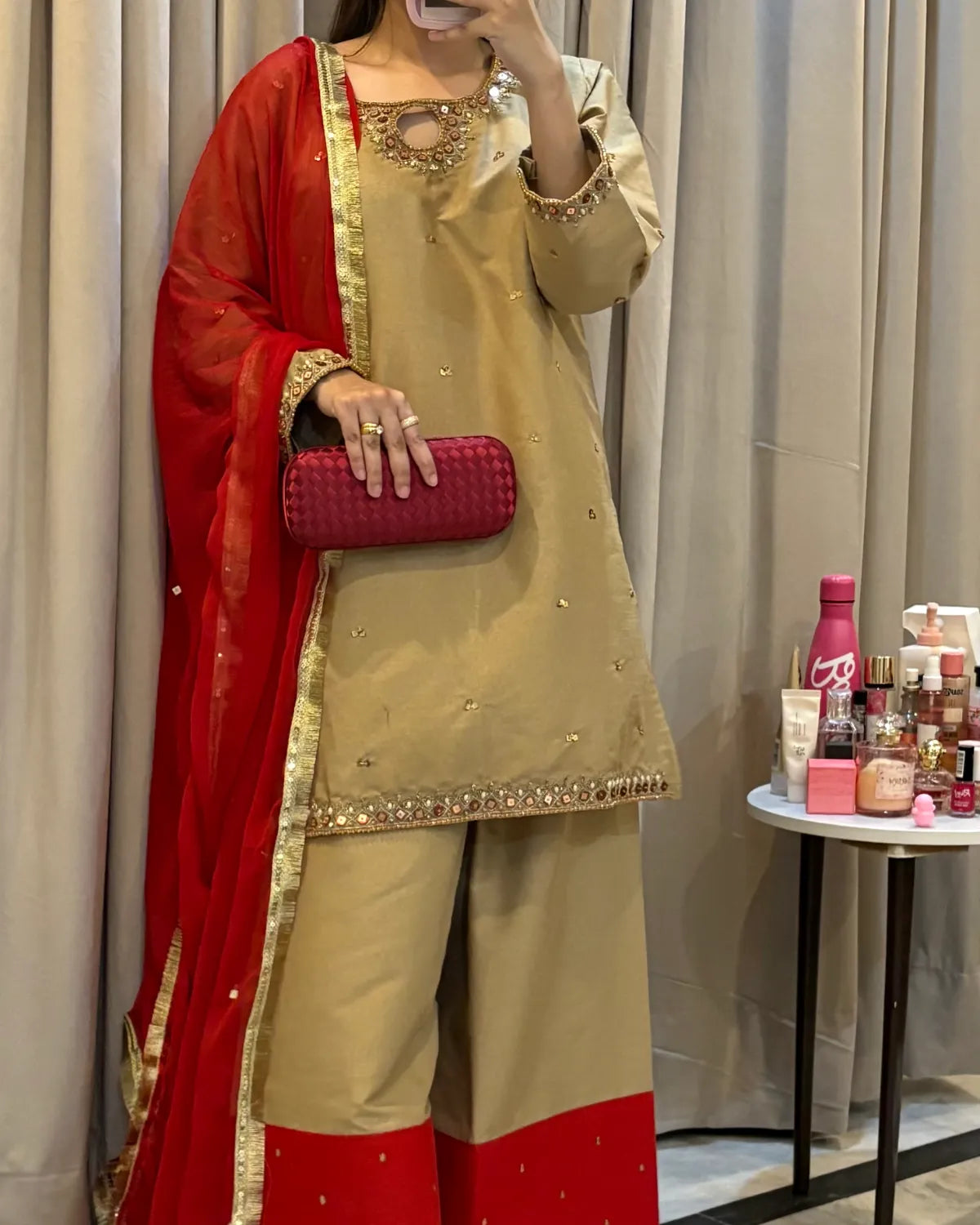A woman models a beige and red traditional South Asian outfit with gold embroidery, holding a red clutch bag. The background features beige curtains and a small side table with cosmetics.