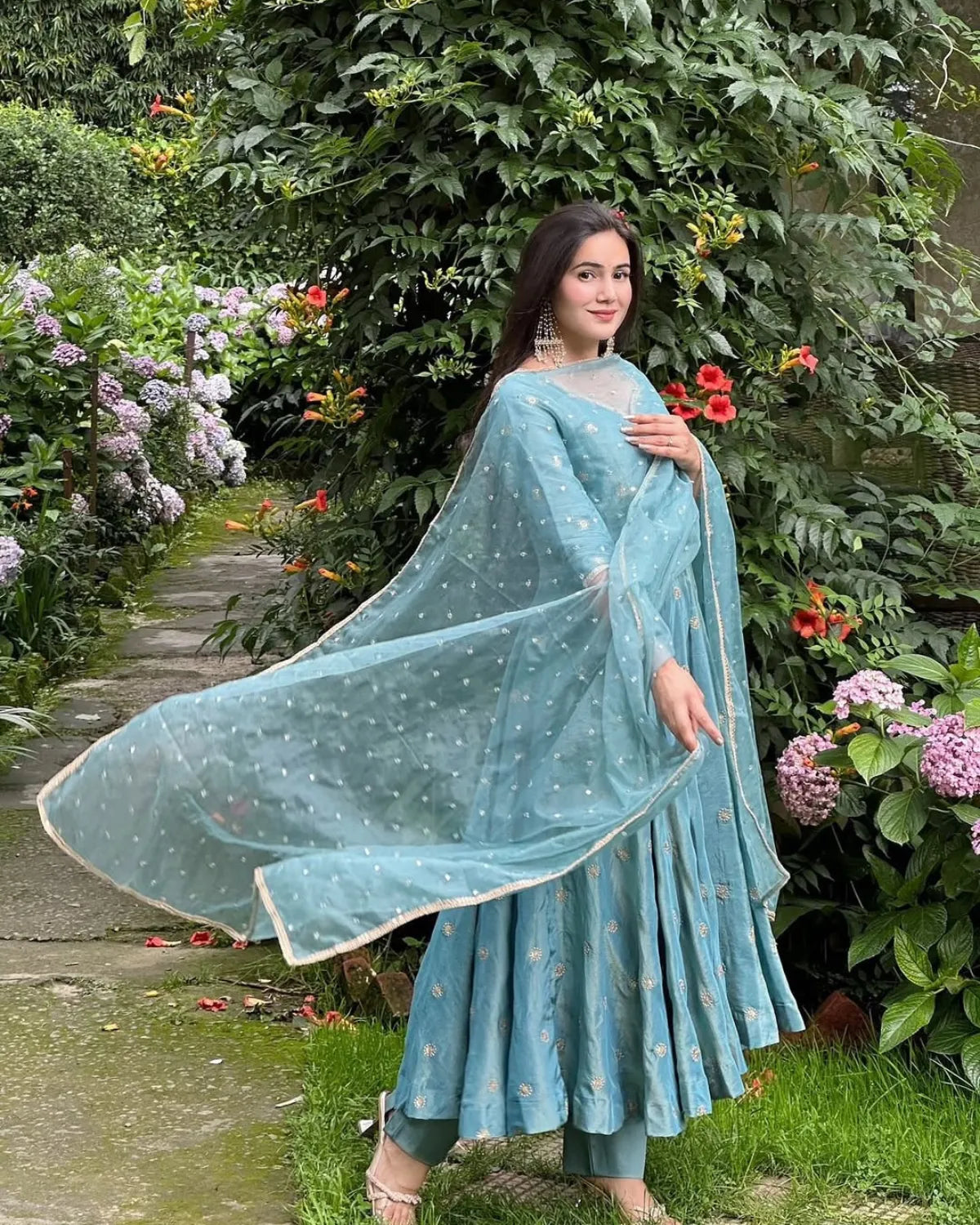 Woman in a light blue traditional outfit standing in a garden with flowers and greenery.
