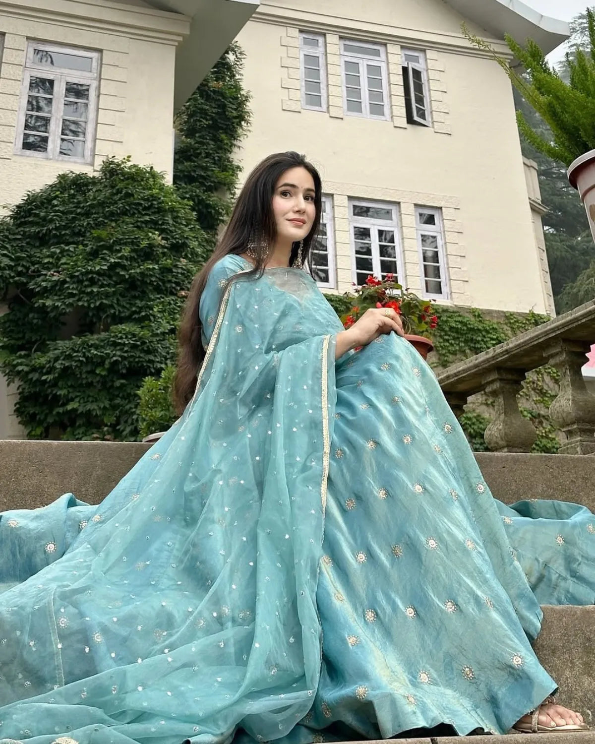 Woman in a light blue saree sitting outdoors with a building in the background