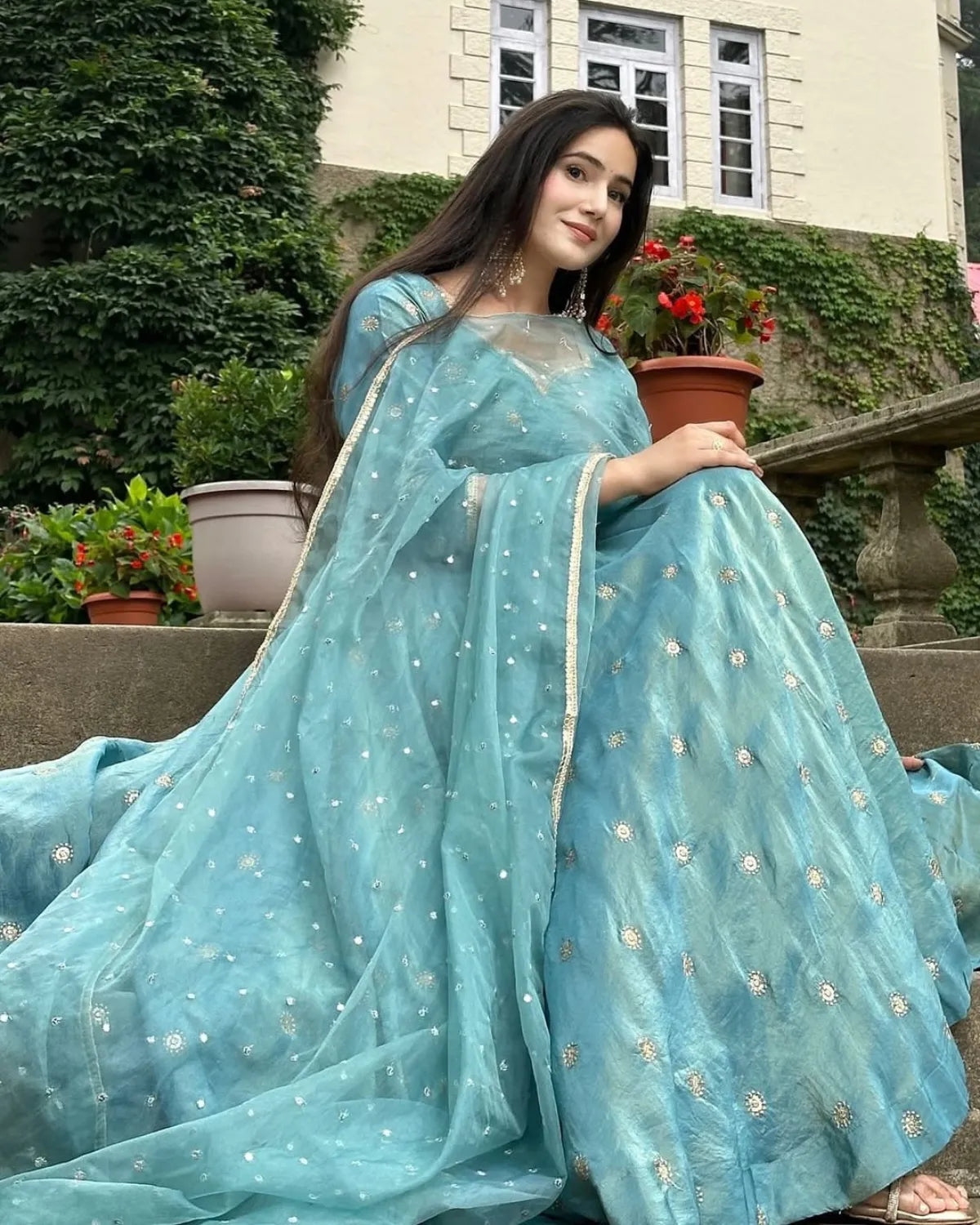 Woman in a light blue saree with floral patterns sitting outdoors.