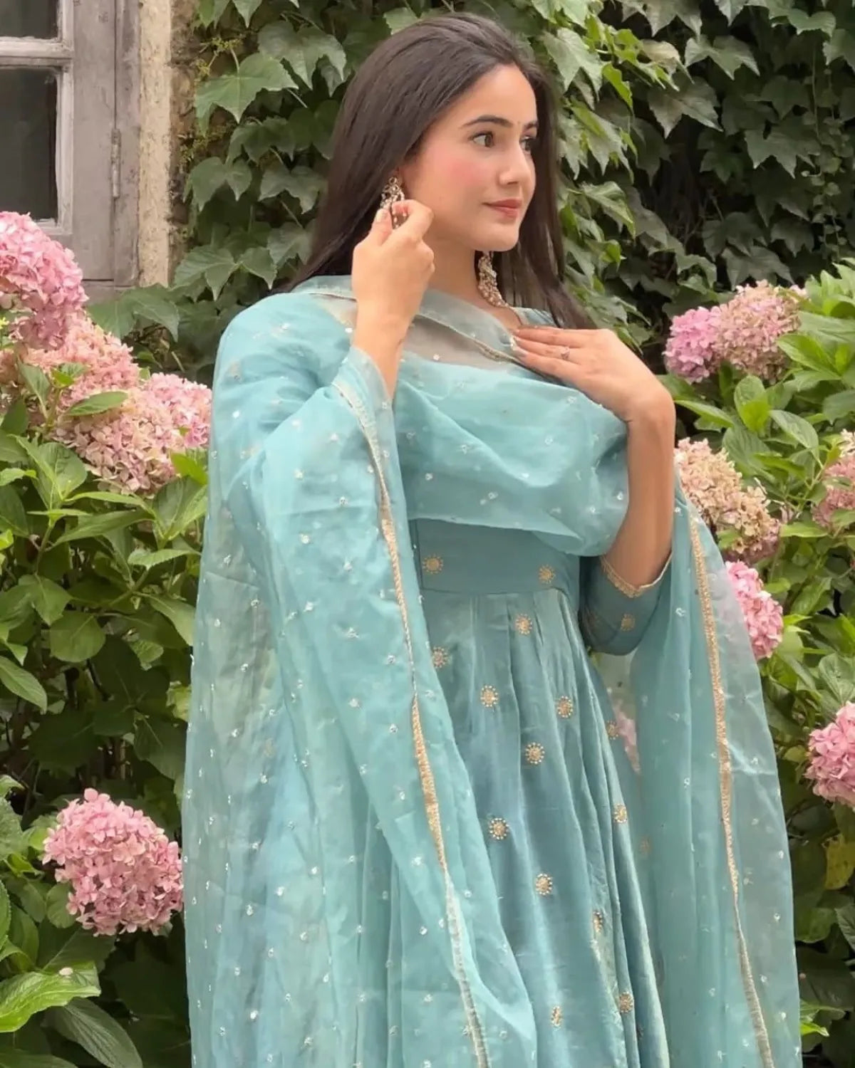 Woman in a light blue saree standing in front of pink flowers and green foliage