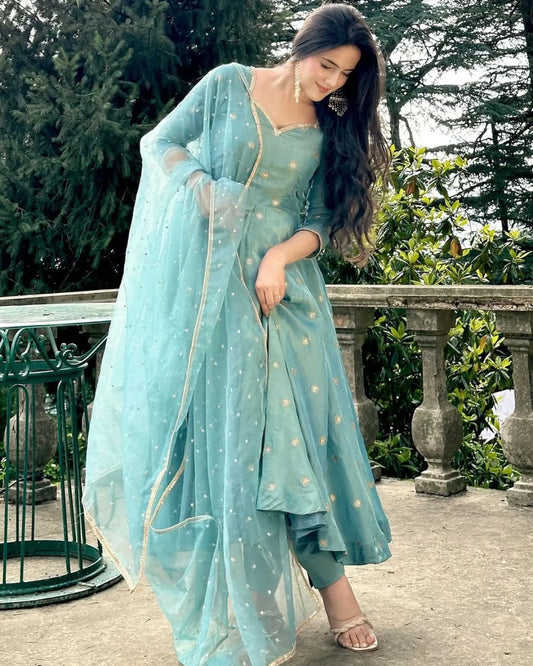 Woman in a light blue traditional outfit standing outdoors with greenery in the background