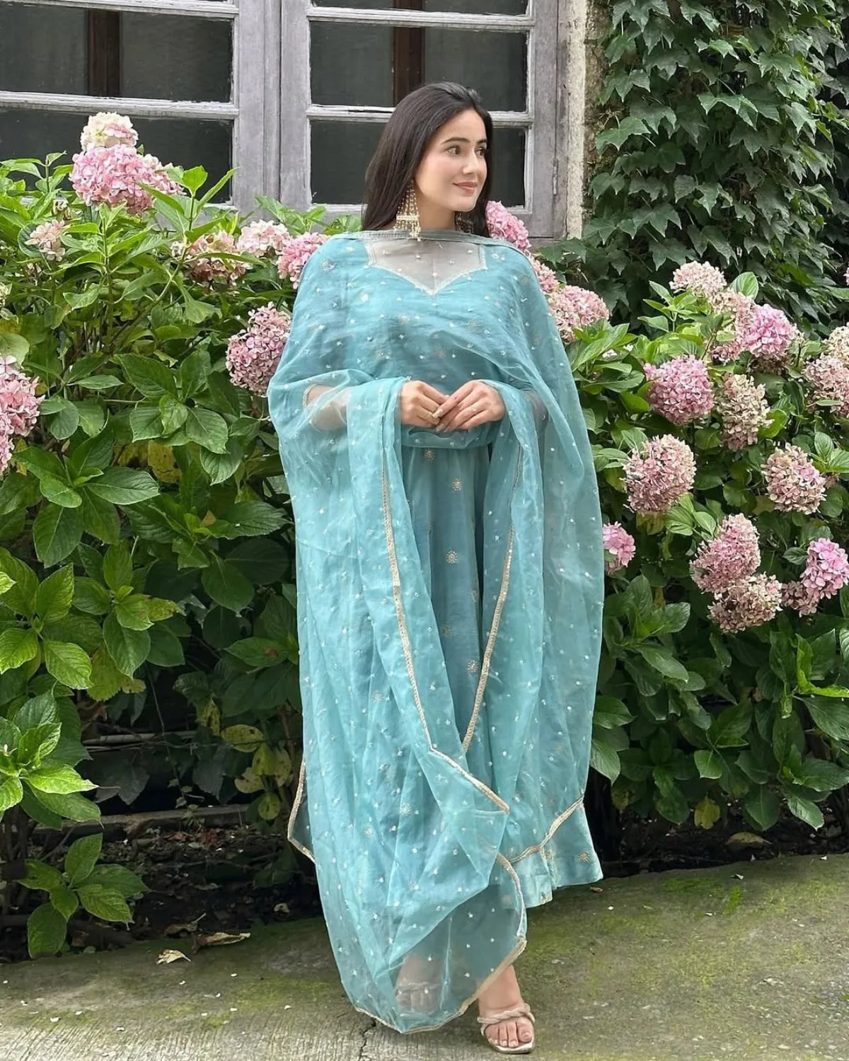 Woman in a light blue traditional outfit standing in front of pink flowers and greenery.