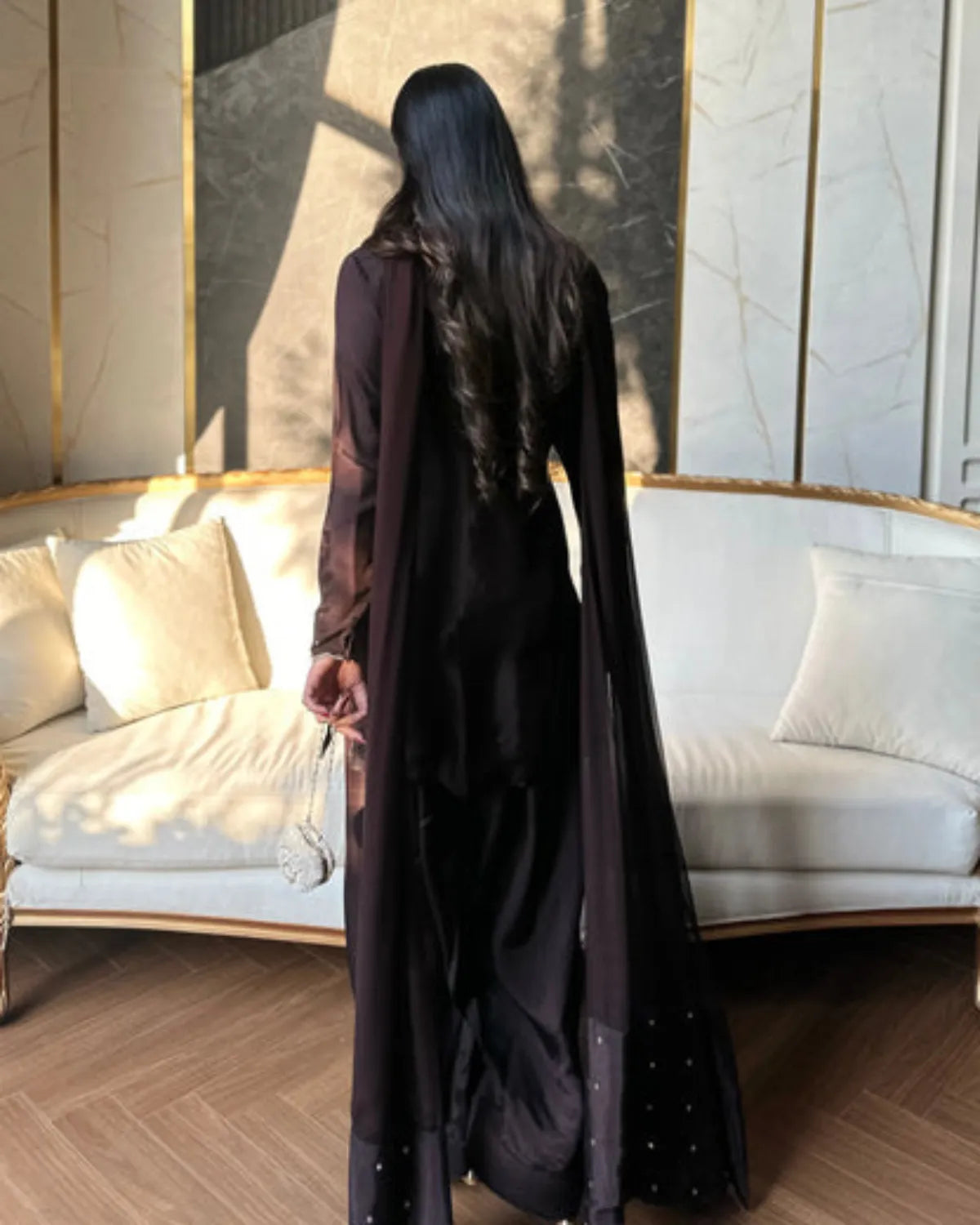 Person wearing a long dark coat standing in a room with a white couch and decorative wall.