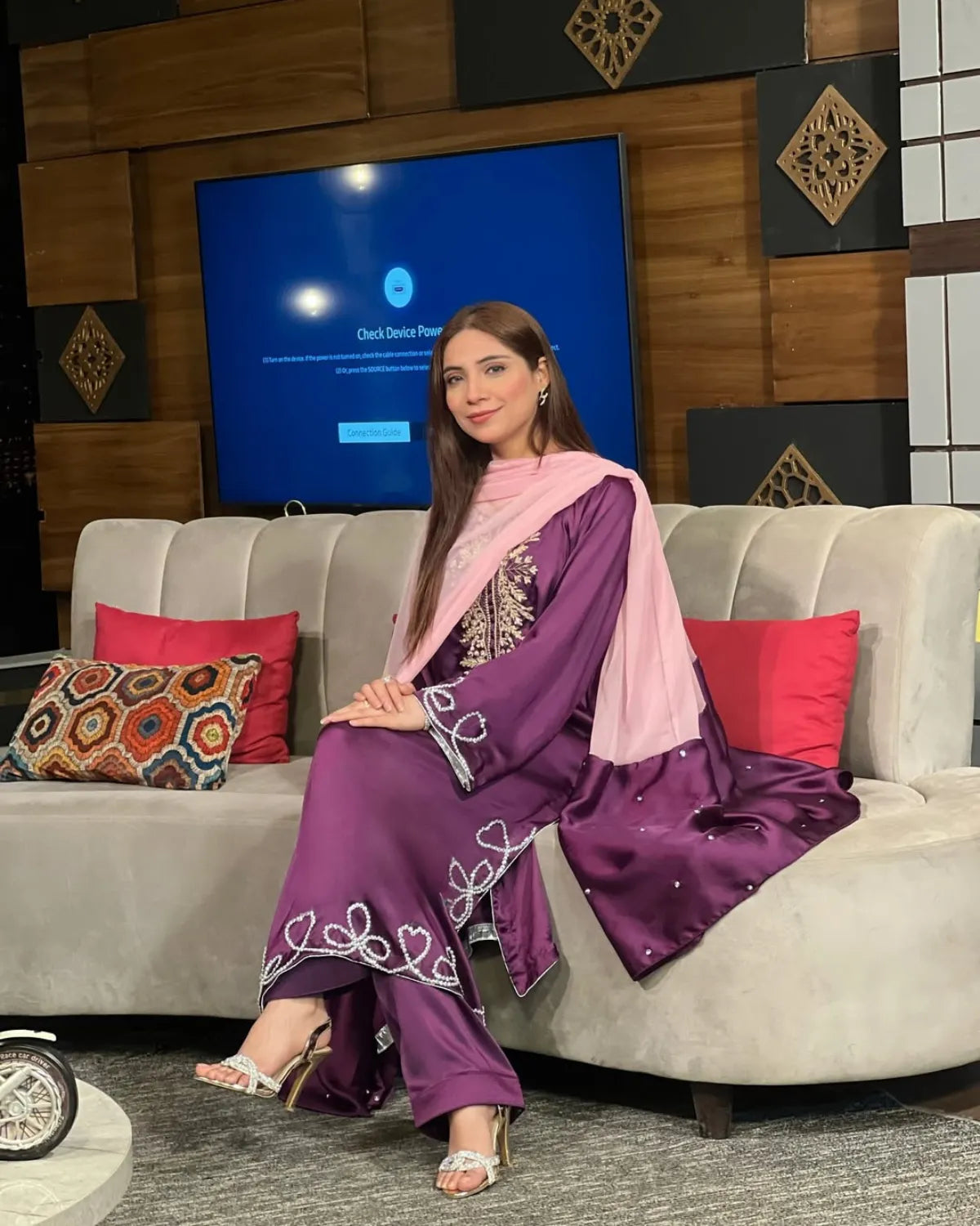 Woman in a purple traditional outfit sitting on a couch with a television screen in the background.