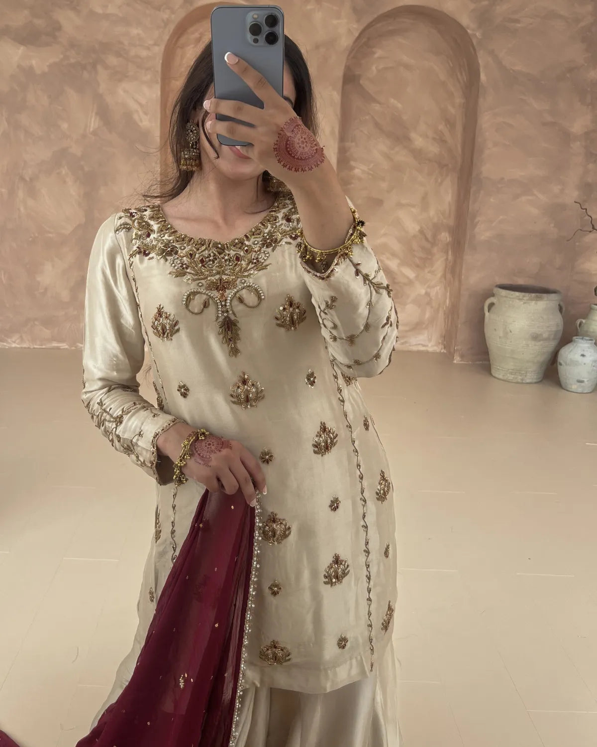Person wearing a cream embroidered outfit with gold details, holding a phone and a maroon fabric.