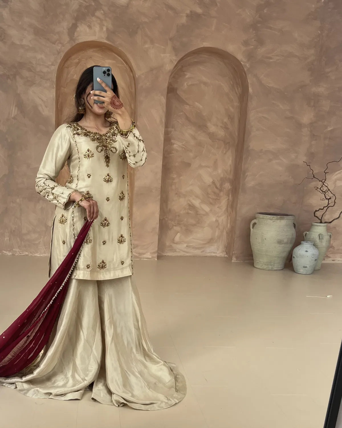 Woman in traditional outfit taking a mirror selfie against a textured wall with decorative vases.