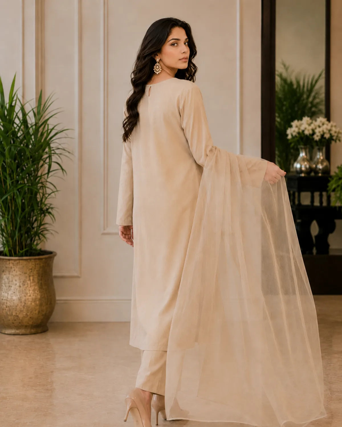 Woman in a beige traditional outfit standing in an elegant room with plants and decor.