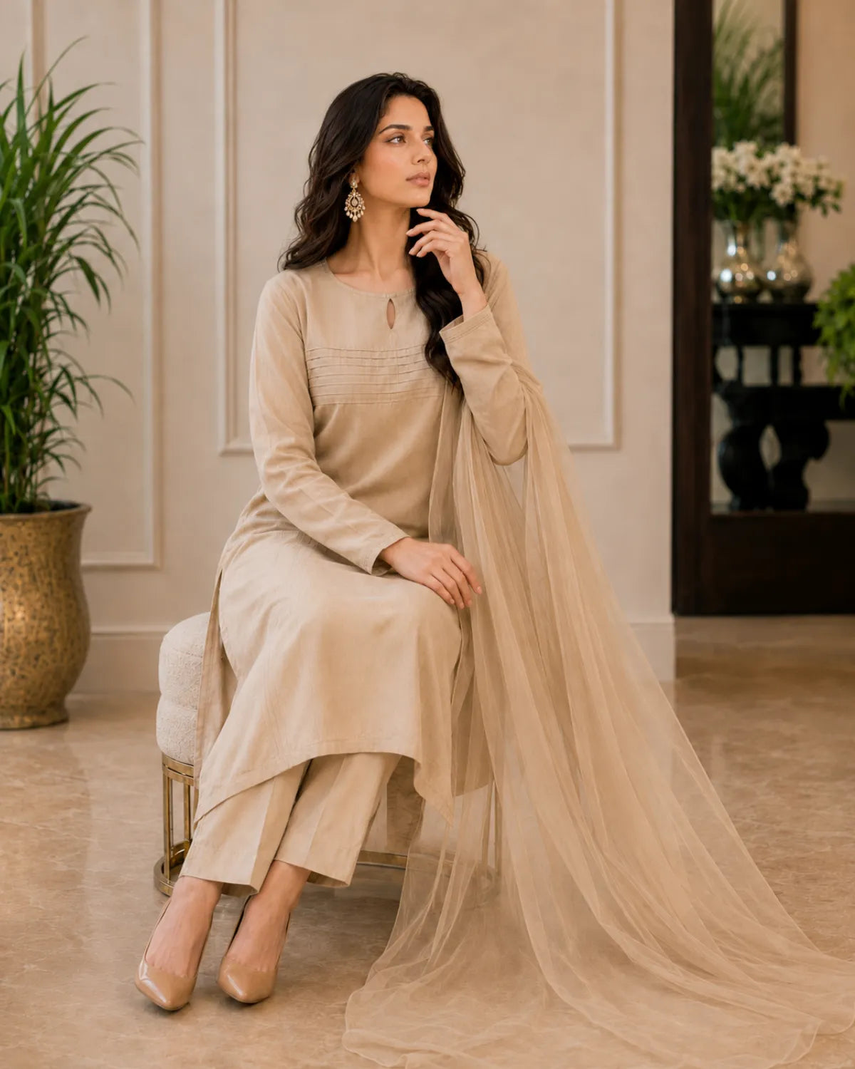 Woman in a beige traditional outfit sitting on a stool in a well-decorated room.