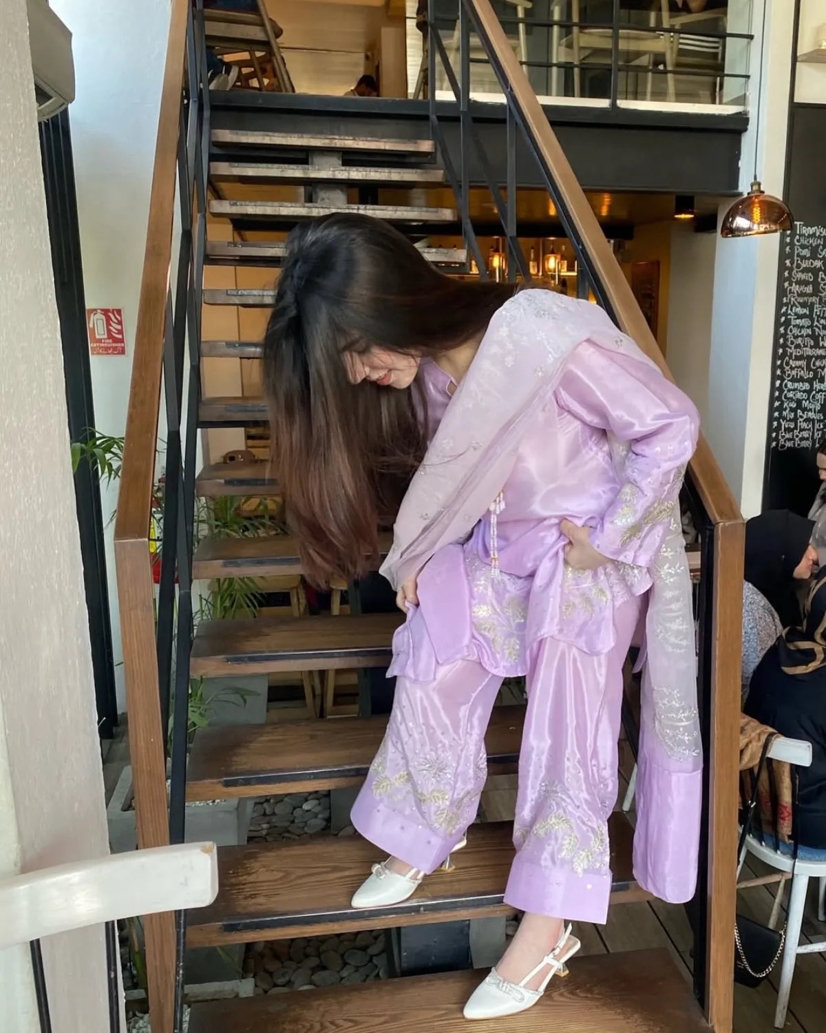 A woman wearing a lavender traditional-style outfit with embroidered details and white pointed shoes, posing on a wooden staircase in a cafe setting.
