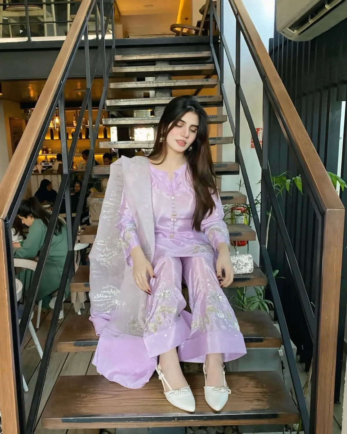 A woman wearing a lavender traditional outfit with a matching sheer dupatta and white pointed shoes, sitting on wooden stairs in an indoor setting.