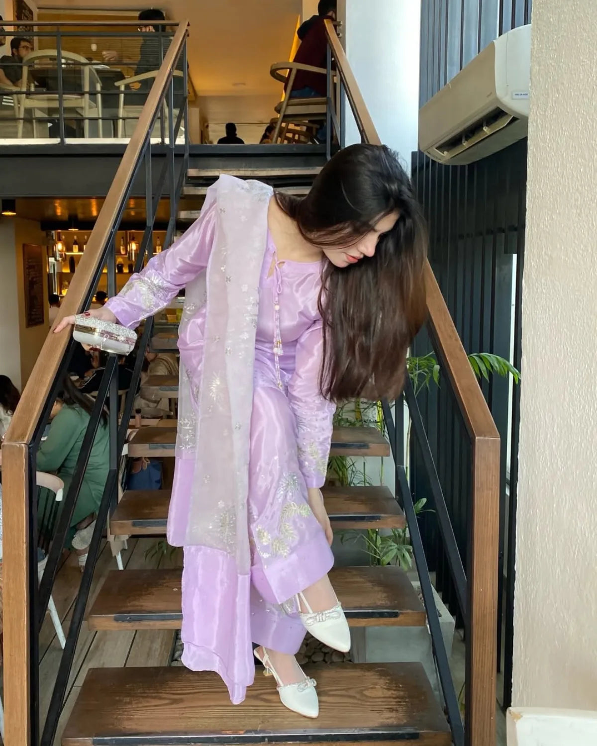 A woman wearing a lavender purple traditional outfit with a matching sheer dupatta and white pointed shoes, posing on a wooden staircase in a cafe setting.
