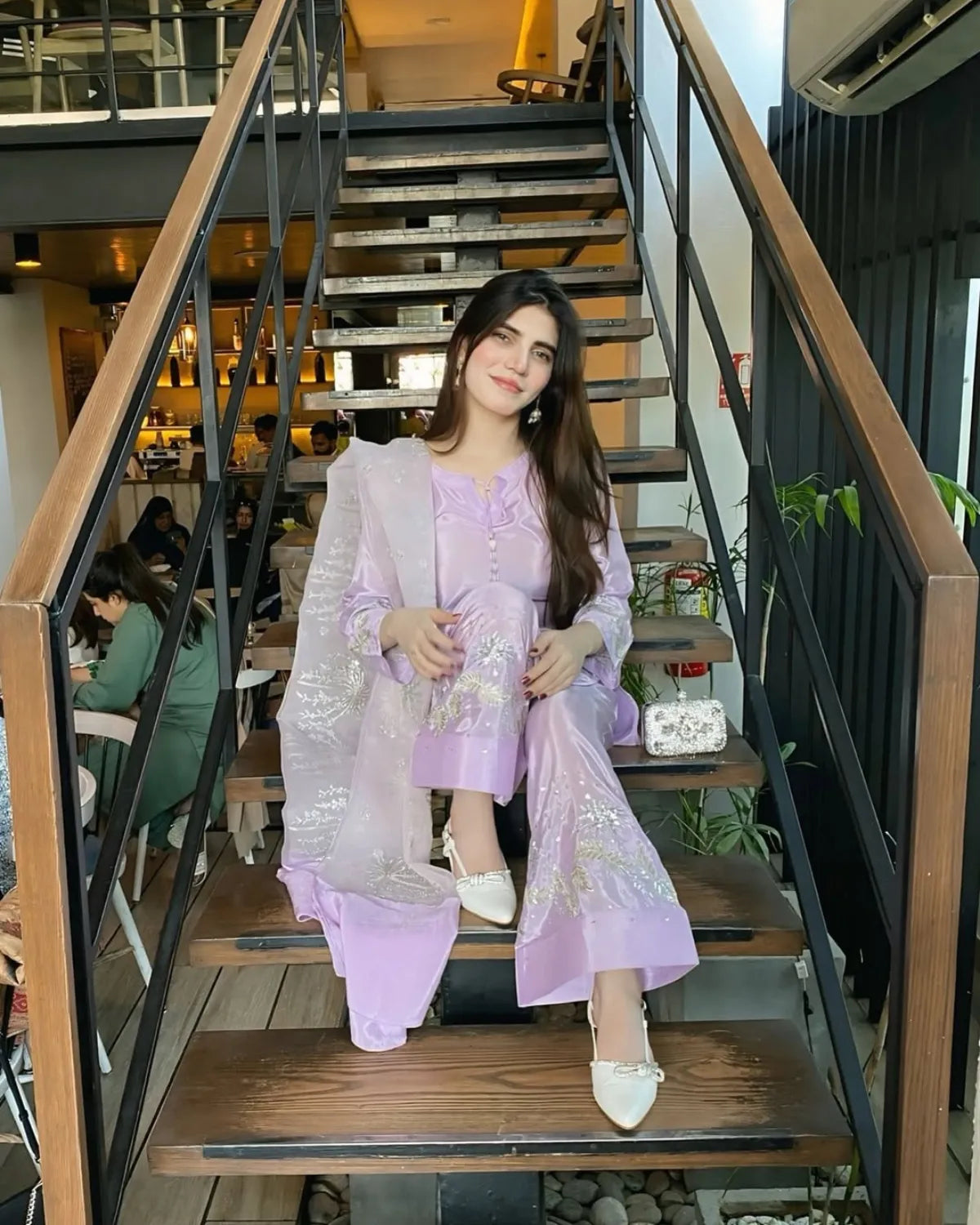 A woman wearing a lavender traditional outfit with a sheer embroidered outer layer sits on wooden stairs in a cafe setting.