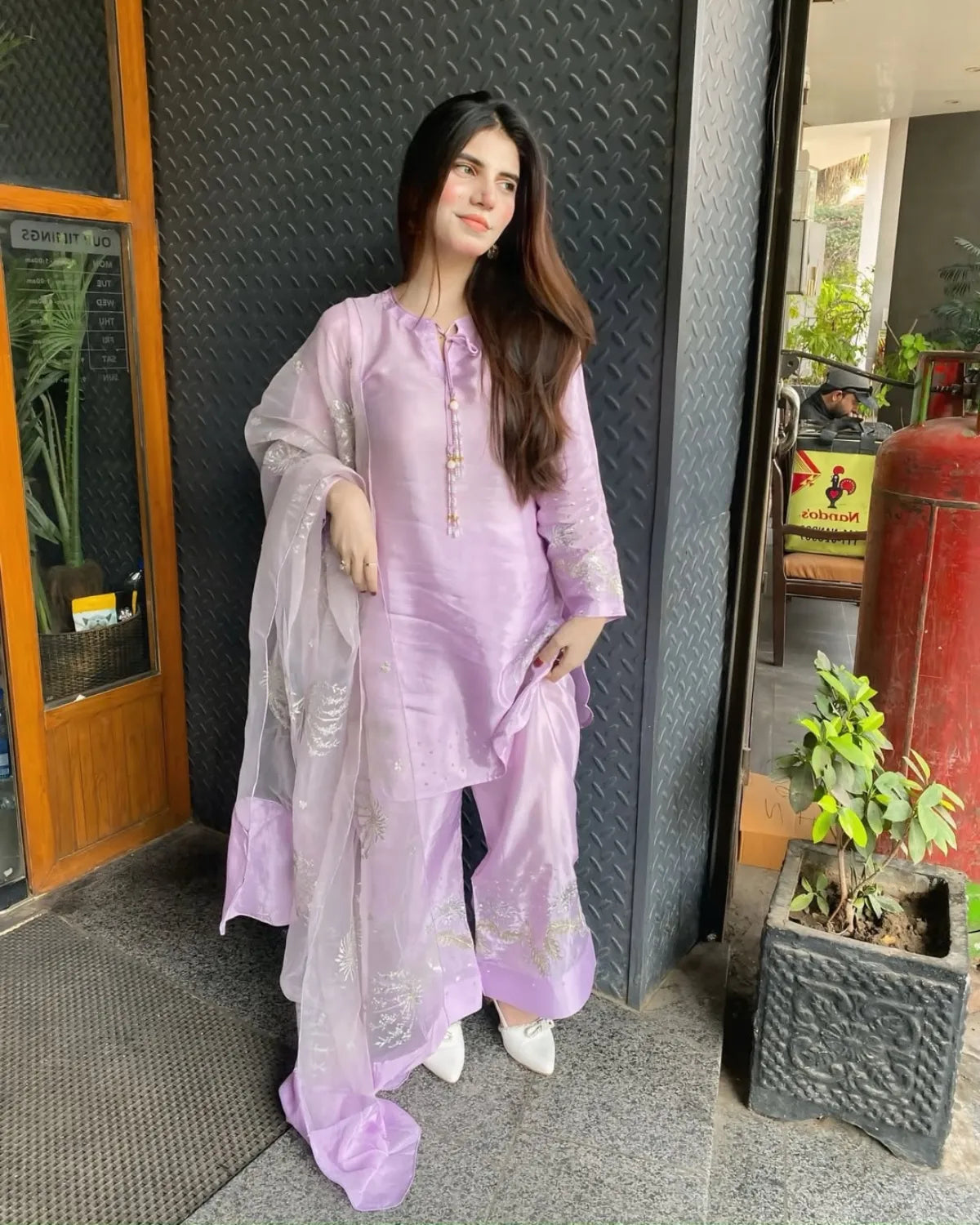 A woman modeling a light purple/lavender traditional three-piece salwar kameez set with a matching sheer dupatta, standing in front of a textured dark gray wall and doorway.