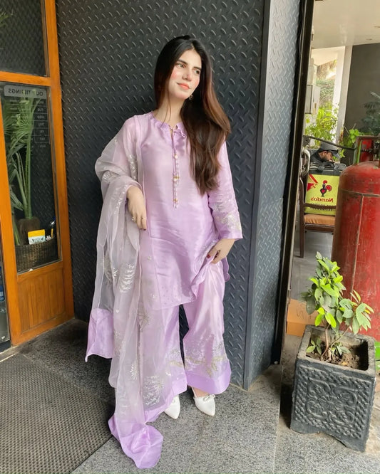A woman modeling a light purple/lavender traditional three-piece salwar kameez set with a matching sheer dupatta, standing in front of a textured dark gray wall and doorway.