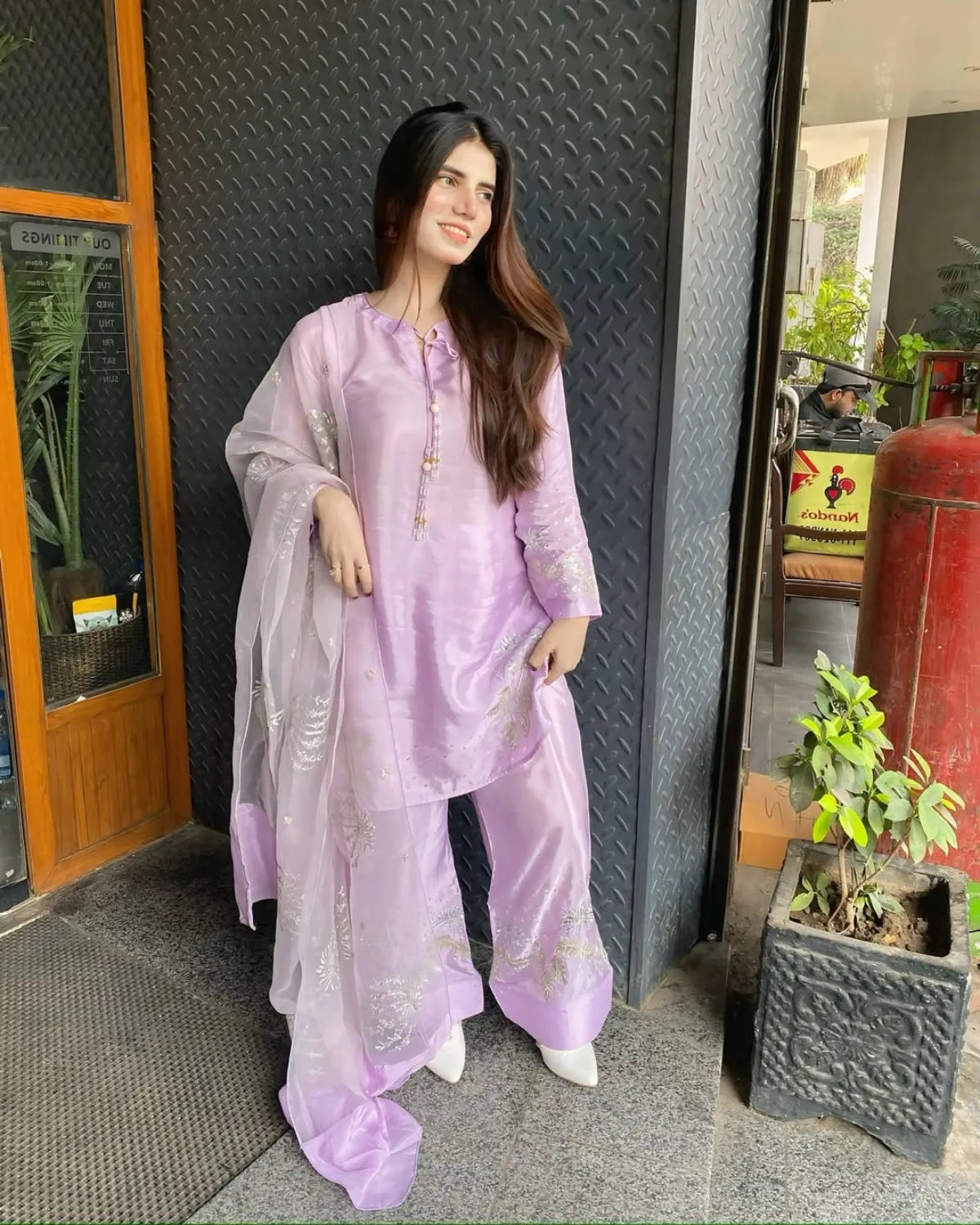 A woman wearing a light purple/lavender three-piece traditional outfit with a matching sheer dupatta, standing in front of a dark textured metal wall and doorway.