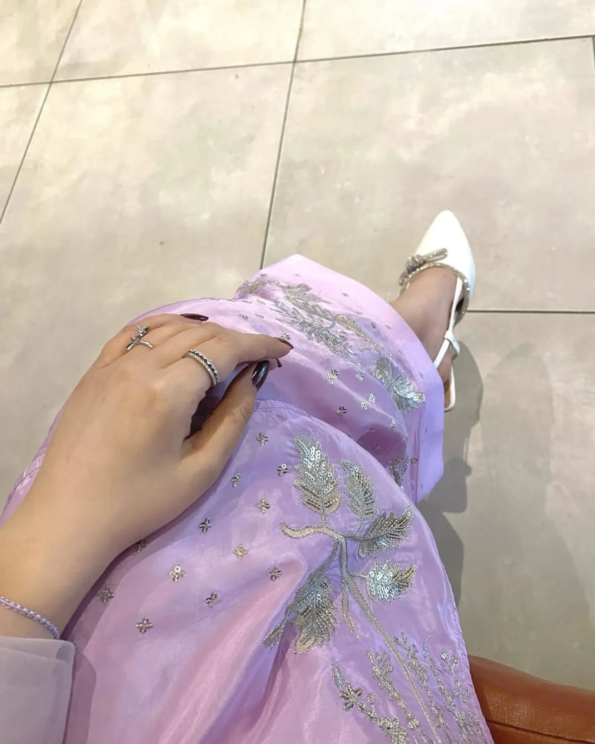 A close-up lifestyle shot of a person wearing a lavender purple tulle skirt with silver and gold floral embroidery, paired with white pointed-toe flats, set against a light beige tiled floor.